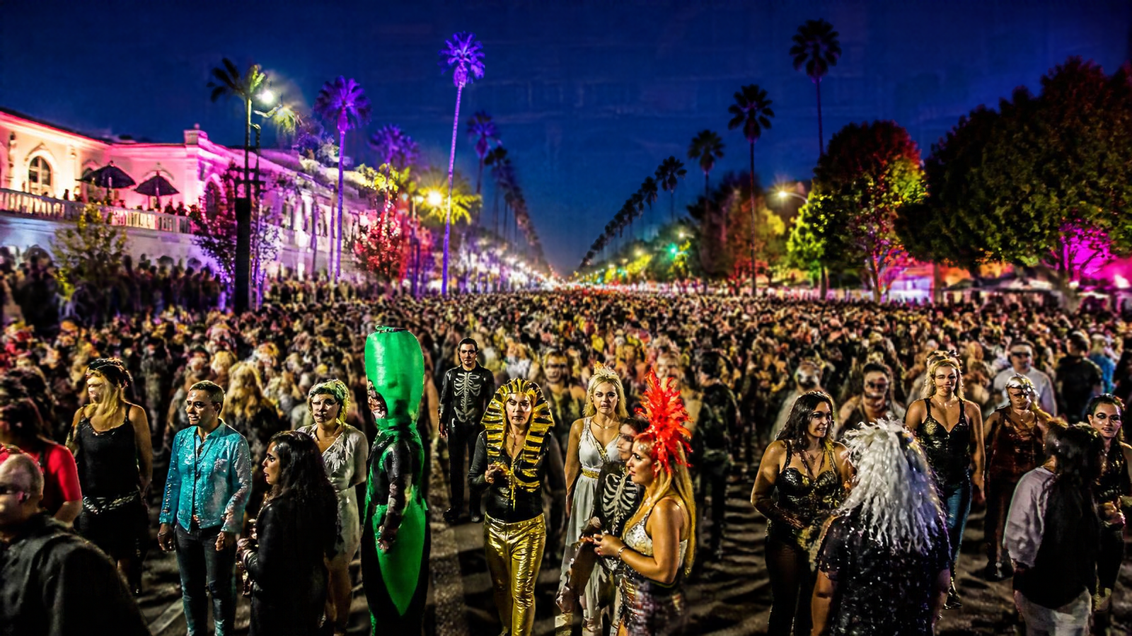 Crowds in colorful Halloween costumes on a neon-lit boulevard lined with palm trees at the West Hollywood Halloween Carnaval